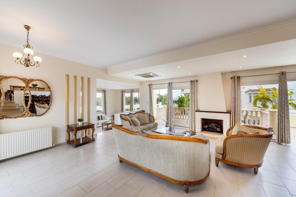A spacious living room with a large sofa, a fireplace, and a view of the outdoors. - Protaras Posidon By The Sea (Paralimni)