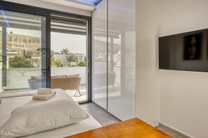 A modern bedroom with a large sliding glass door leading to a balcony, a bed with white linens, a wicker chair, and a flat-screen TV mounted on the wall. - Lux Deck Roof Apt at the Foothills of Acropolis w Breathtaking View (Athens)