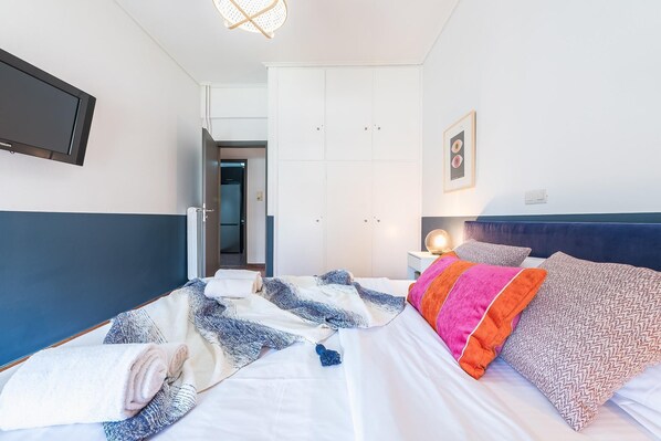 A hotel room with a bed, a bedside table with a lamp, a wall-mounted TV, and a door leading to another room.