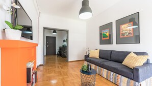 A modern living room with a sofa, a coffee table, and a TV mounted on the wall. There are framed pictures on the wall and a potted plant on the floor. - New Relaxed Central Apartment Walk Everywhere (Athens)