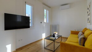 A living room with a flat-screen TV mounted on the wall, a yellow sofa, a coffee table with a book and a bowl, and a radiator. - Paradise Suite With Jacuzzi Acropolis View (Athens)