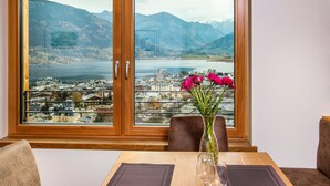 Un coin repas avec une table dressée pour deux personnes, un vase rempli de fleurs et une vue sur un lac et des montagnes, visible à travers une grande fenêtre.