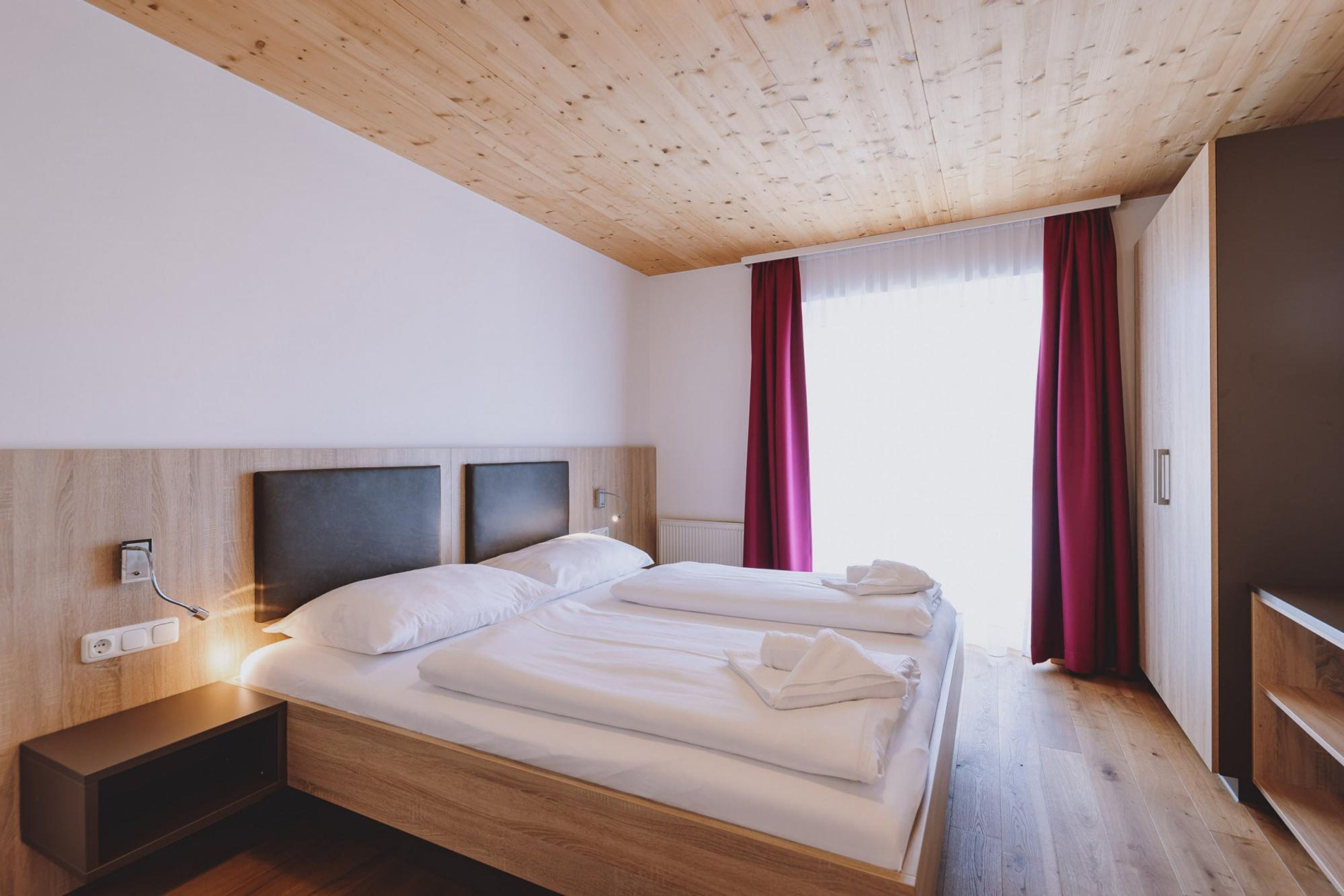 A modern hotel room with a wooden ceiling, a large bed with white linens, a bedside table, and a view of the outdoors through a window with red curtains.