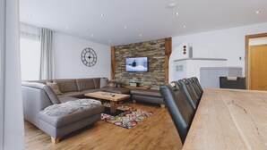 Ein modernes Wohnzimmer mit Ecksofa, Couchtisch aus Holz, an einer Steinwand montiertem Fernseher und Essbereich mit Holztisch und Stühlen.