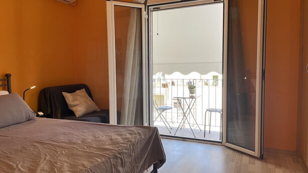 Un dormitorio con cama, una silla, un balcón con mesa y sillas, y vistas al exterior.