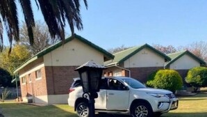 A barbecue grill with sausages and fruits, a parked car, and a house in the background. - Twin Room in Hillside - 2087 (Bulawayo)