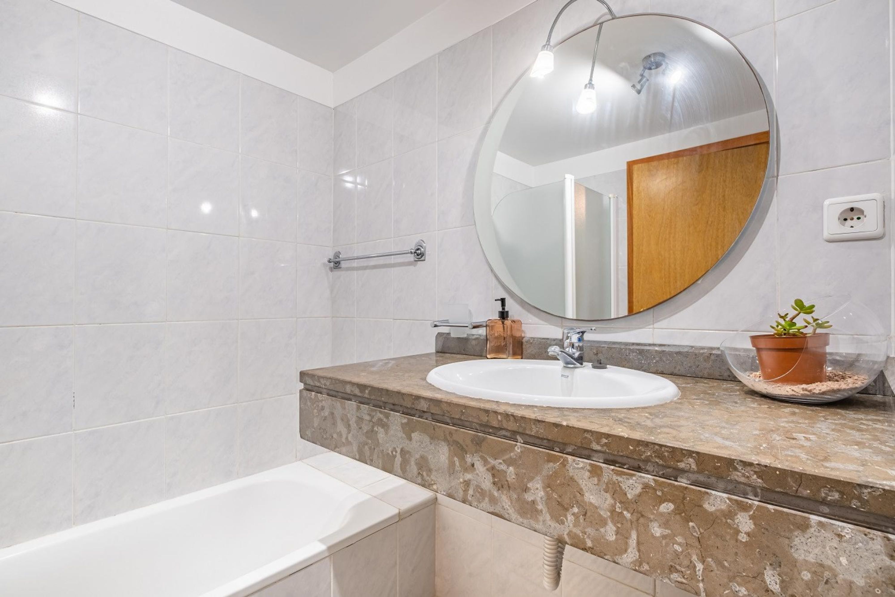 A modern bathroom with a white bathtub, a round mirror, a sink with a soap dispenser, and a potted plant on the countertop.