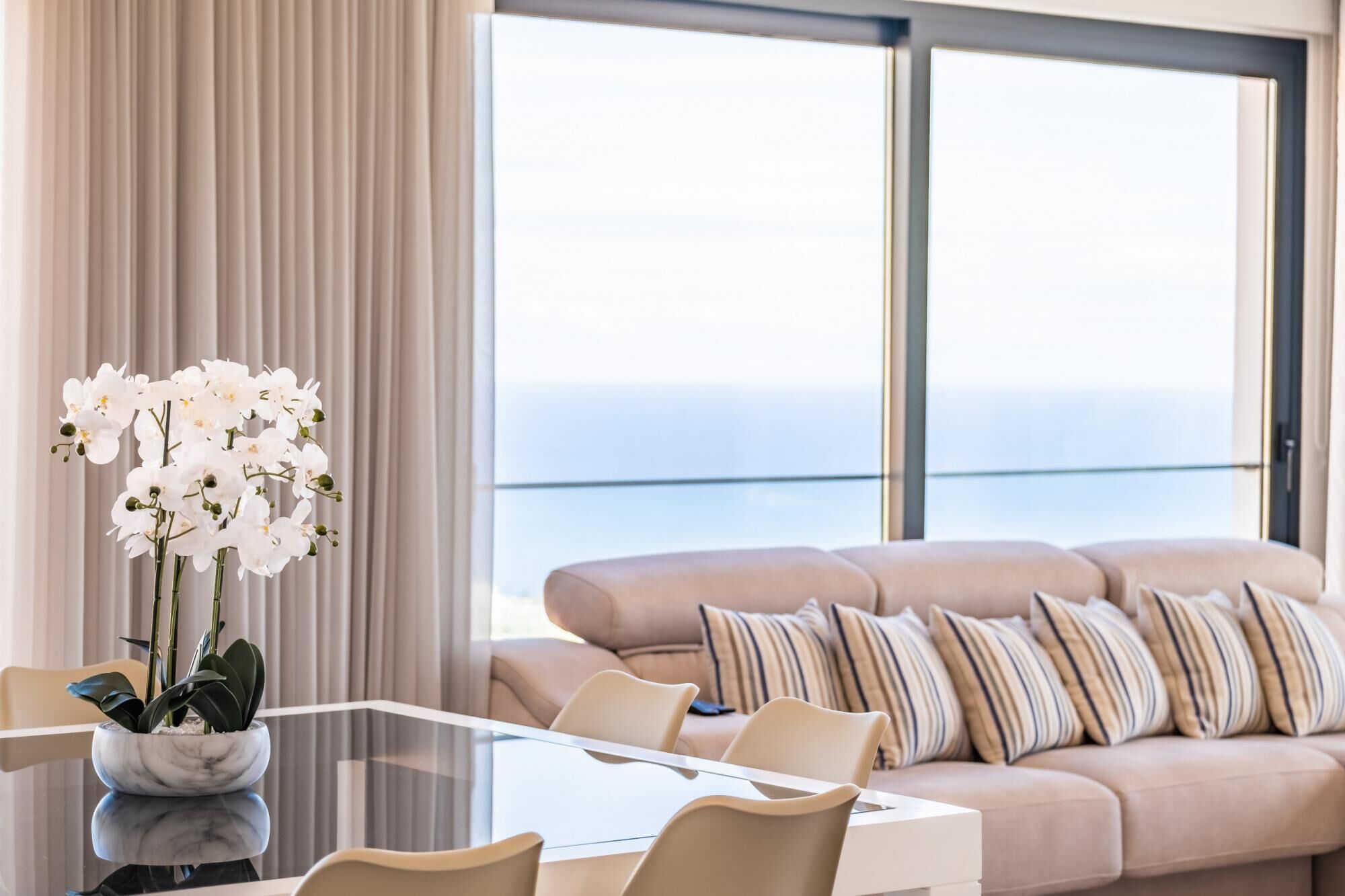 A modern living room with a glass table, beige sofa, and white orchids.