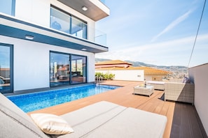 Un área de piscina exterior moderna con cubierta de madera, sillones de descanso y vistas a la ciudad y colinas al fondo.