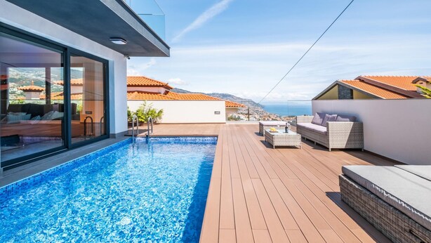 A modern outdoor pool area with a wooden deck, a glass-enclosed lounge, and a view of the sea and cityscape.