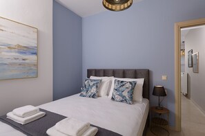 Una cama bien hecha con sábanas blancas y almohadas de flores azules, una lámpara de noche y un cuadro en la pared.