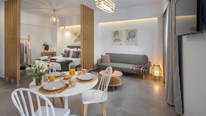 A modern living room with a sofa, dining table, and chairs. There is a bed with a headboard and a small table with a lamp. The room has a neutral color scheme and wooden accents. - Myra Mare White Suite by Estia (Heraklion)