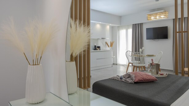 A modern hotel room with a glass coffee table, a bed with a dark bedspread, a bedside table with a pink pillow, a large mirror, and a view of the dining area. - Myra Mare White Suite by Estia (Heraklion)