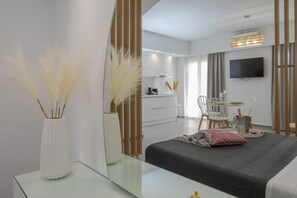 A modern hotel room with a glass coffee table, a bed with a dark bedspread, a bedside table with a pink pillow, a large mirror, and a view of the dining area. - Myra Mare White Suite by Estia (Heraklion)