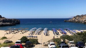 Una playa con hileras de sombrillas y tumbonas, un litoral rocoso y un cielo azul despejado.