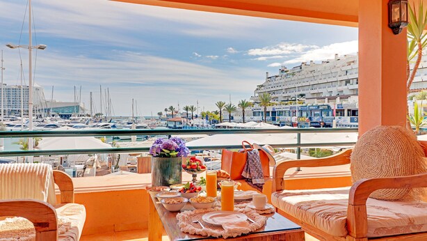 A balcony with a view of a marina, featuring a table with breakfast items, a vase of flowers, and a wicker chair. - Garden Marina in Vilamoura (Loulé)