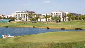 A golf course with a water body, surrounded by modern buildings with multiple floors and large windows. - Garden Marina in Vilamoura (Loulé)