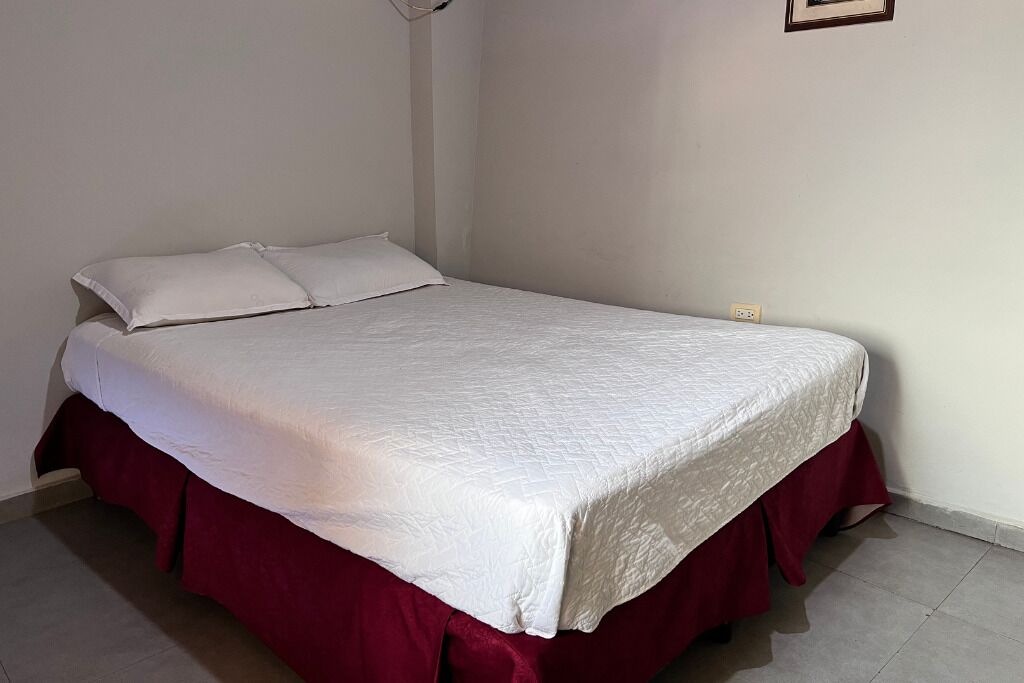 Een eenpersoonsbed met een witte geborduurde spreidekker en rode poten met een rok, in een kamer met effen witte muren en een betegelde vloer.