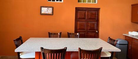 A dining area with a rectangular table, six wooden chairs, a wooden door, and a window with curtains.