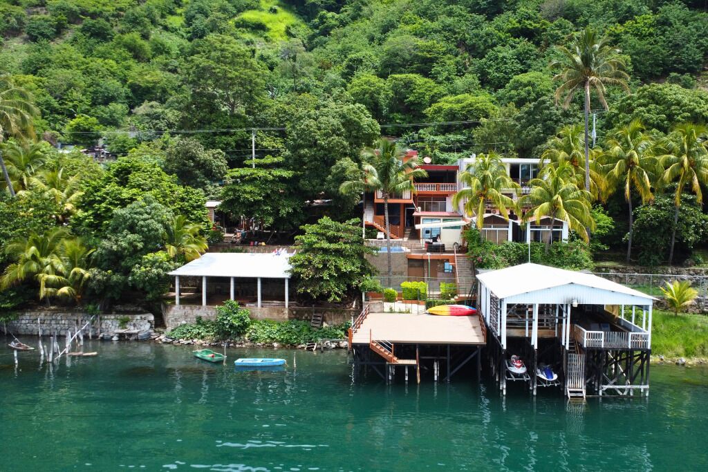 Une maison construite sur pilotis, au-dessus de l’eau, entourée d’une végétation luxuriante et de palmiers.
