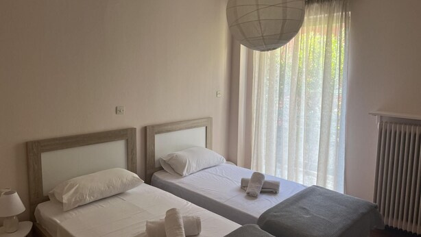 A hotel room with two single beds, a bedside table with a lamp, a large window with sheer curtains, and a radiator.
