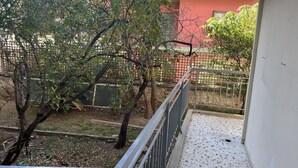 A balcony with a metal railing overlooking a garden area with trees and a red building in the background.