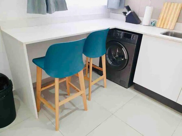 A compact kitchen with a washing machine, two blue chairs, and a sink.