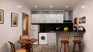 A compact kitchen with white cabinets, a washing machine, a sink, and a fruit bowl on the counter. There are two wooden chairs and a round table. - Vila do Largo A by Homie (São Vicente)