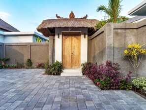 Traditional entrance with a thatched roof, wooden door, and stone wall, surrounded by lush greenery.