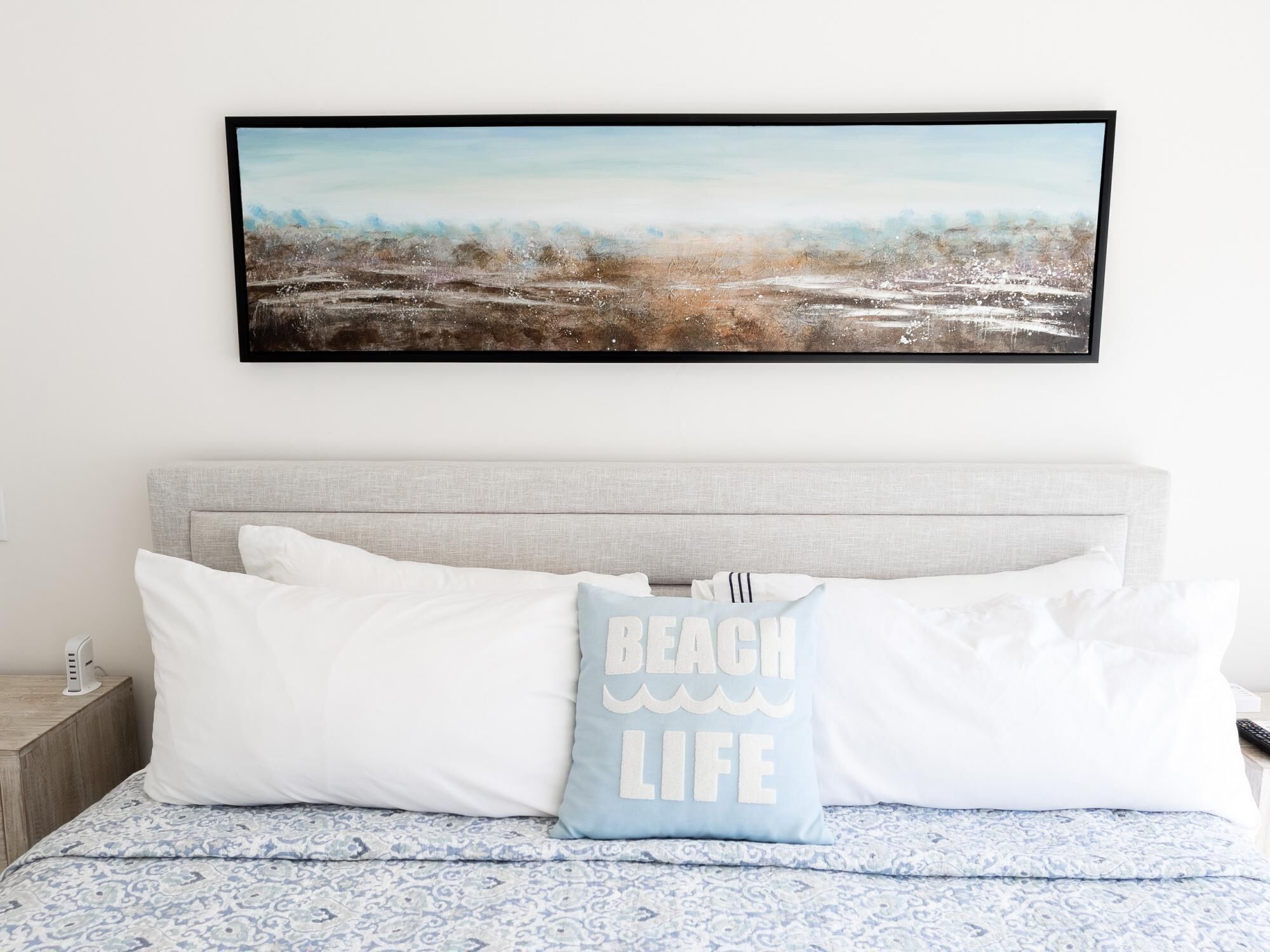 A bedroom with a bed, white pillows, a blue and white patterned bedspread, and a framed beach-themed artwork on the wall.