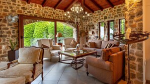 A spacious living room with stone walls, a chandelier, and a view of greenery outside. - Luxury Villa Golden Crest With Private Swimming Pool (Hersonissos)