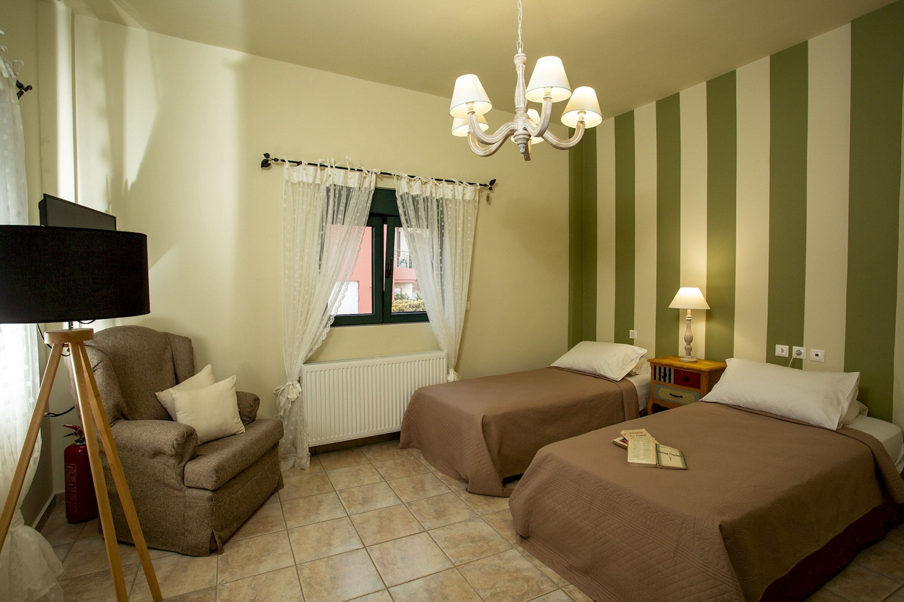 A hotel room with two beds, a chair, a lamp, a window with curtains, and a chandelier.
