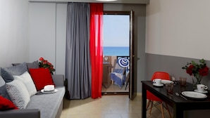 Un salon moderne avec un canapé gris, des coussins rouges et un coin repas avec une table dressée pour deux personnes. Une grande fenêtre offre une vue sur la mer.