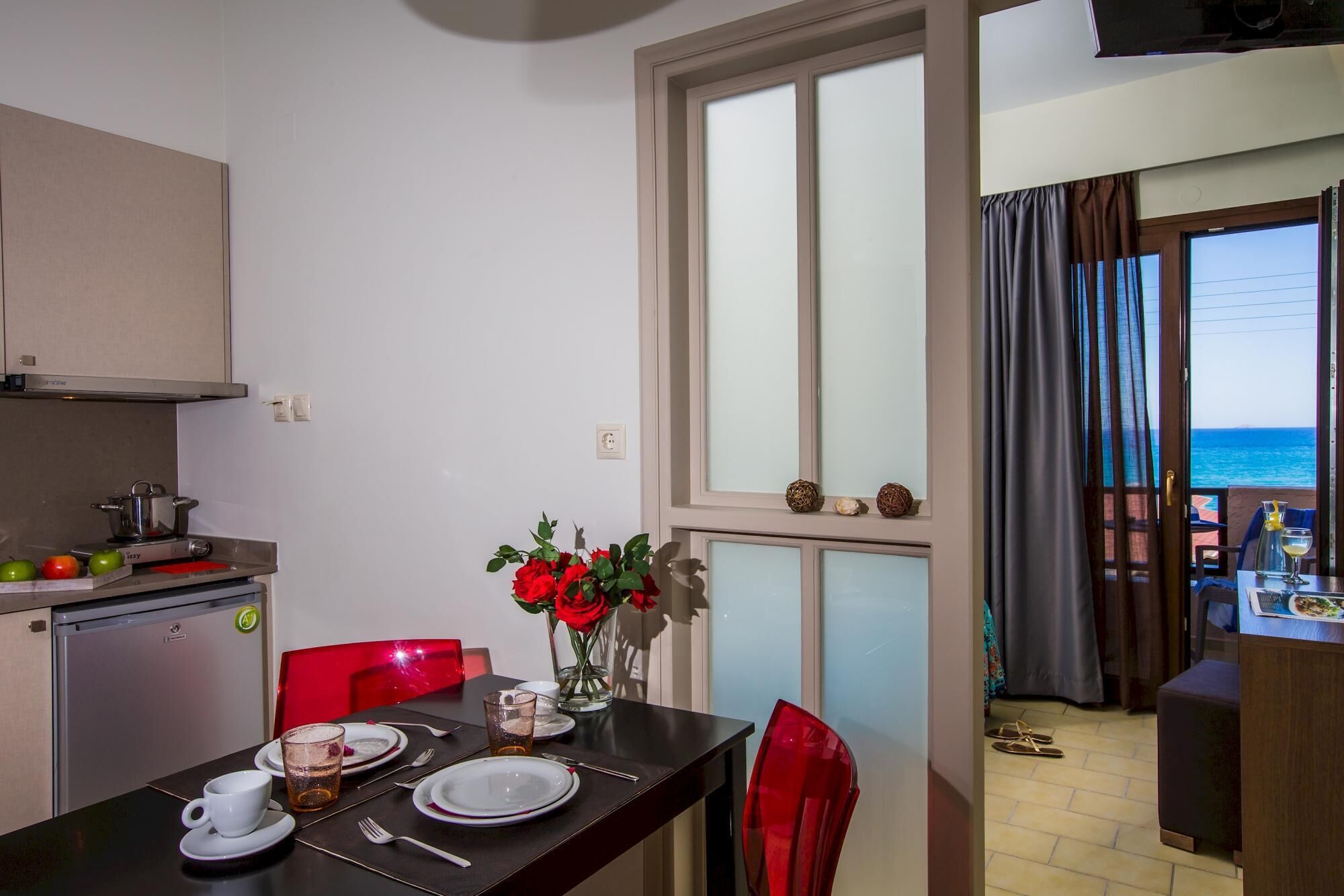 A dining area with a table set for two, a kitchen area with a refrigerator and a stove, and a view of the sea through a window.