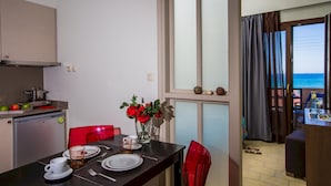 Un coin repas avec une table dressée pour deux personnes, un espace cuisine équipé d’un réfrigérateur et d’une cuisinière, et une vue sur la mer par une fenêtre.