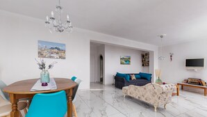 A modern living room with a sofa, a dining table with chairs, a TV, and a chandelier. - Olive Tree Villa by Estia (Corfu)