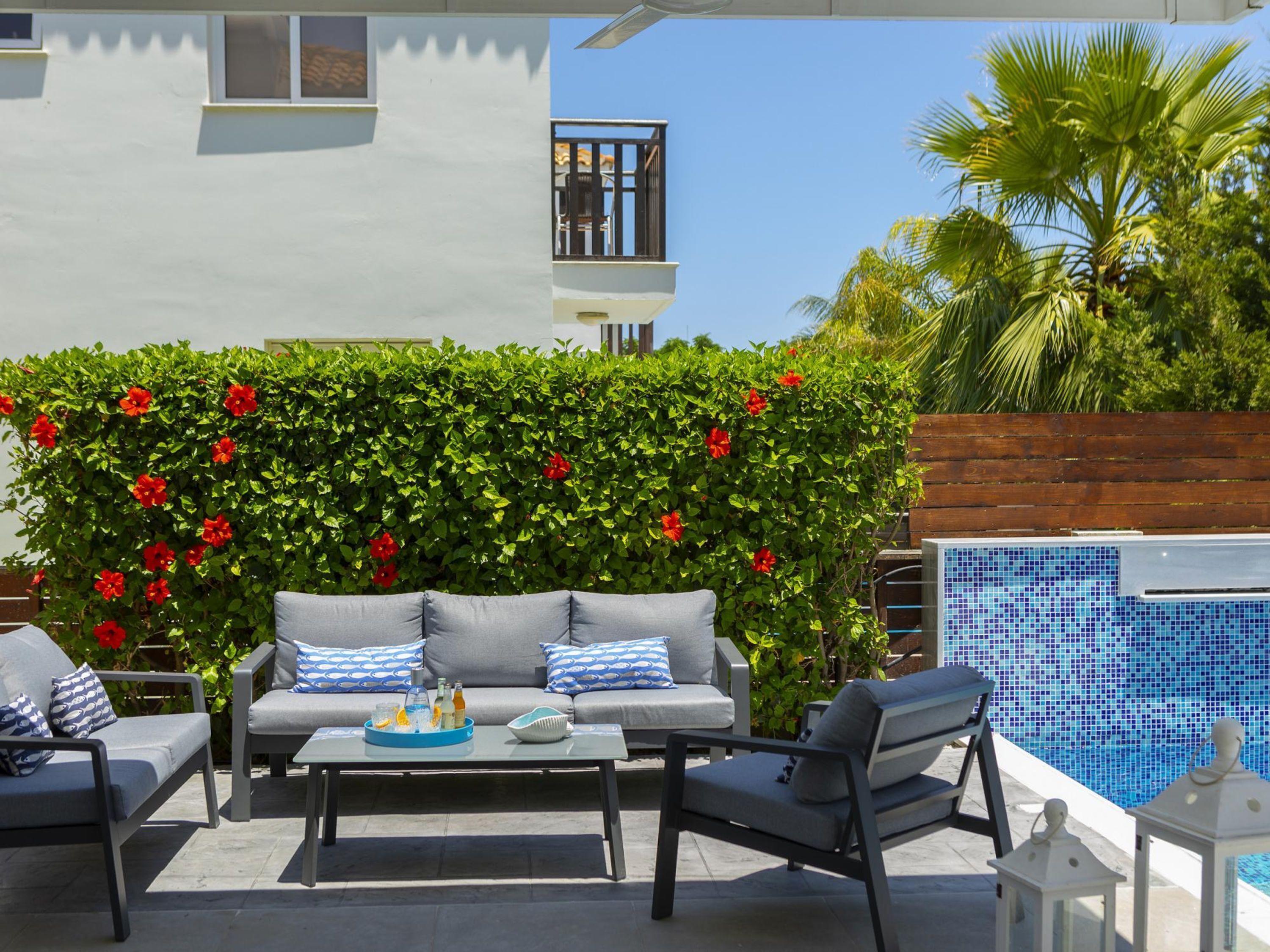 An outdoor seating area with a sofa and chairs, a table with drinks, and a pool in the background.
