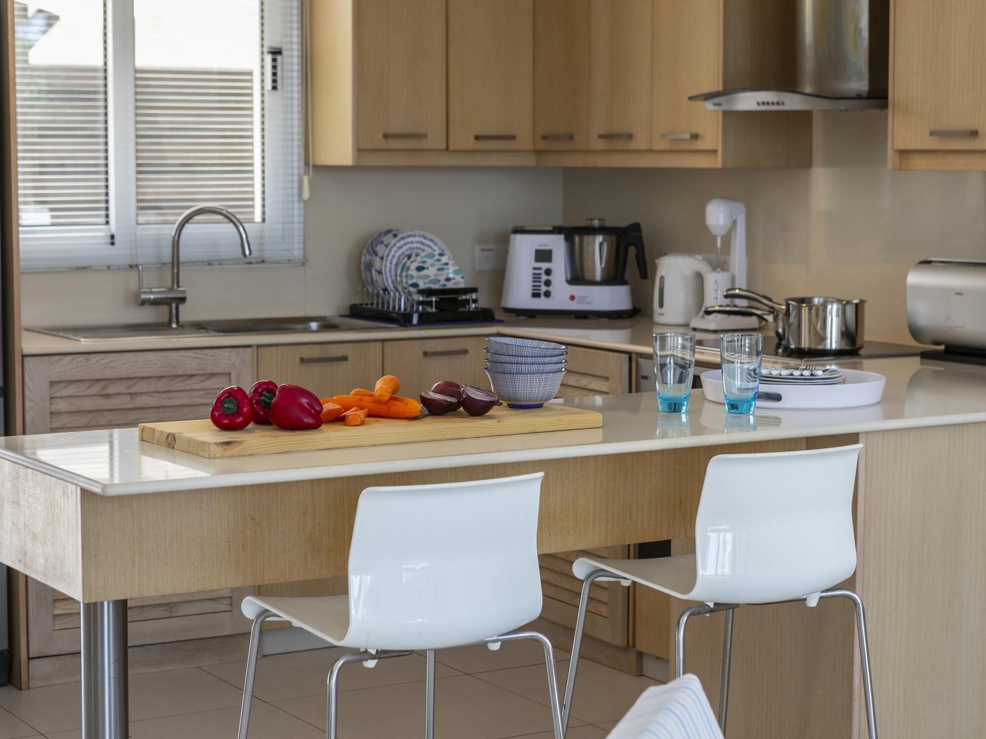 A modern kitchen with wooden cabinets, a central island with a cutting board, and two white chairs.