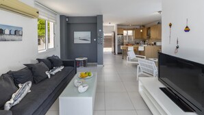 Ein modernes Wohnzimmer mit Sofa, Couchtisch und Fernseher. Im Hintergrund ist eine Küche mit Holzschränken und einem Essbereich zu sehen.