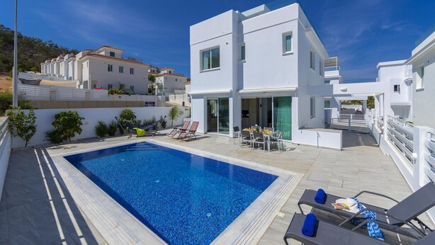 Un edificio moderno de color blanco con piscina, sillas de descanso y zona de comedor.