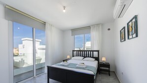 Un dormitorio moderno con una cama grande, una puerta corrediza de vidrio a un balcón, una mesita de noche pequeña con una lámpara y un aire acondicionado instalado en la pared.
