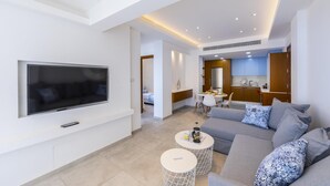 A modern living room with a flat-screen TV, a grey sofa, two round coffee tables, and a kitchen area in the background. - Mackenzie Castella Beachfront Suite (Larnaca)