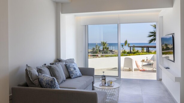 A modern living room with a grey sofa, a coffee table, and a TV on the wall. A large sliding glass door leads to an outdoor area with a view of the beach and palm trees. - Mackenzie Castella Beachfront Suite (Larnaca)