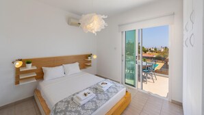 A modern bedroom with a wooden headboard, a large bed with white linens, a hanging light fixture, and a balcony with outdoor furniture. - Ayia Napa Marina Vibes (Sotira)