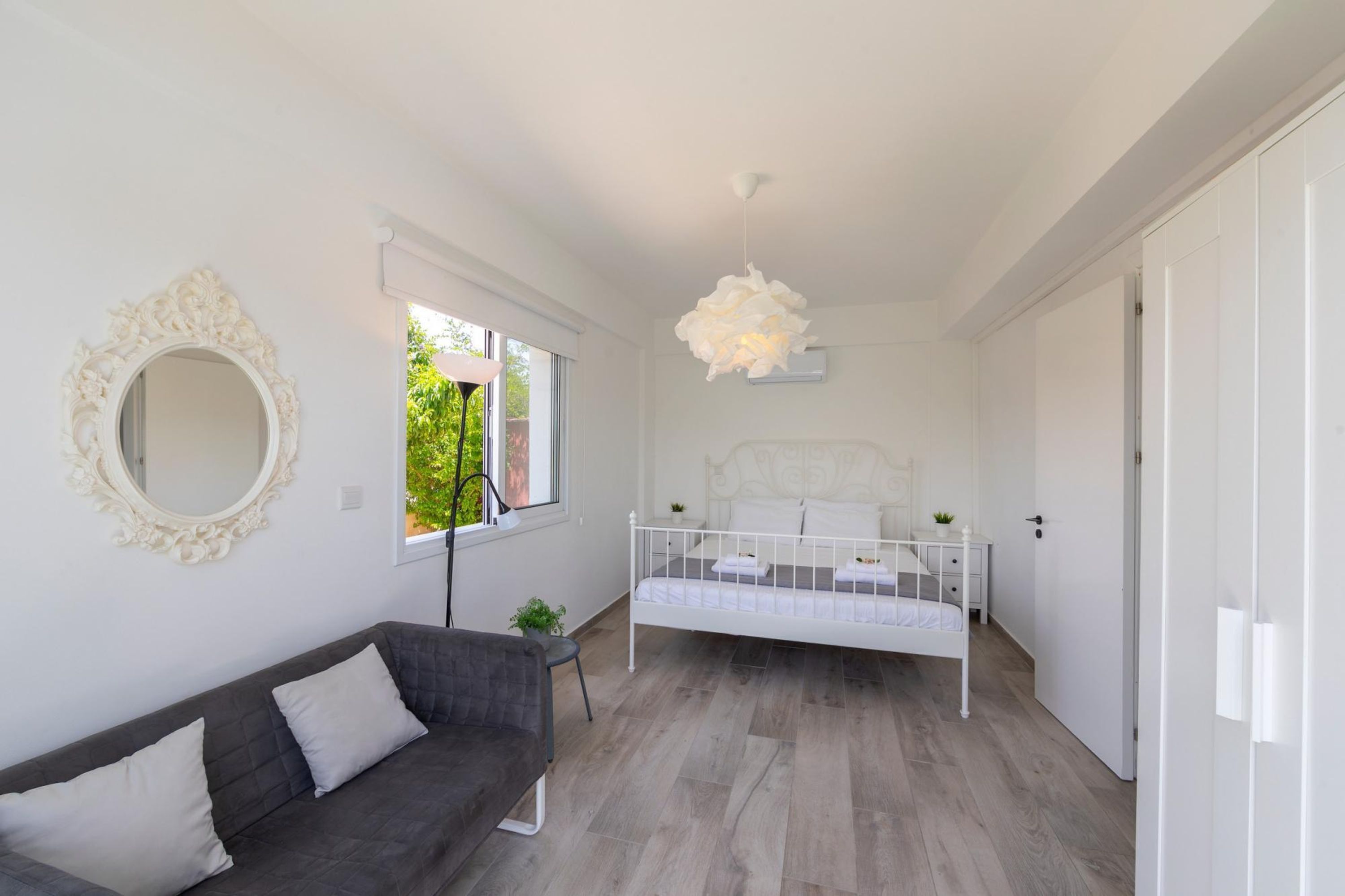 A modern bedroom with a white bed, a gray sofa, a large mirror, and a hanging light fixture.