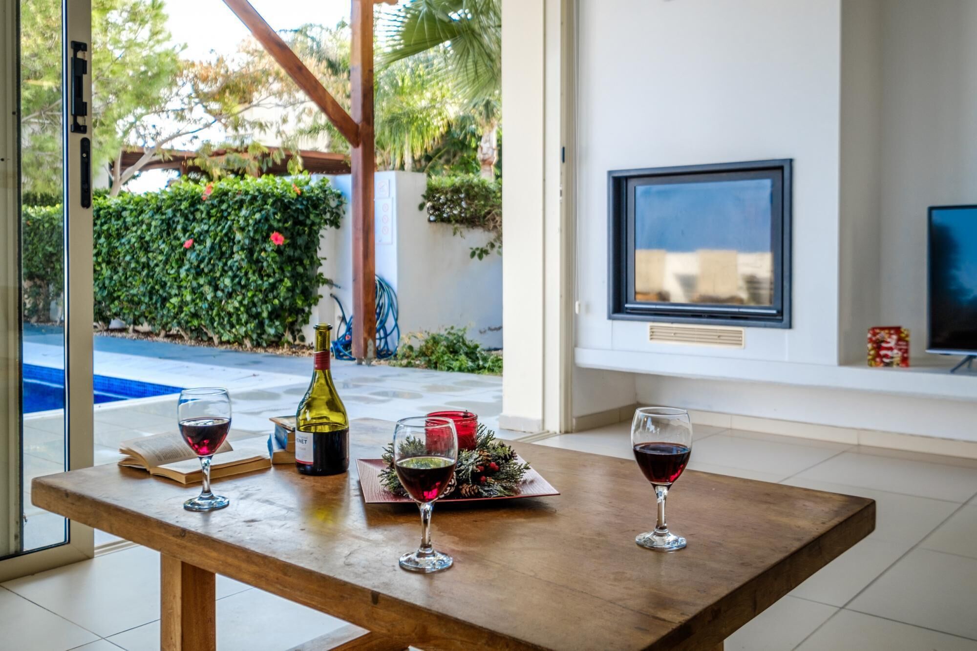 A table with wine glasses and a bottle, a framed picture on the wall, and a view of a pool and greenery outside.