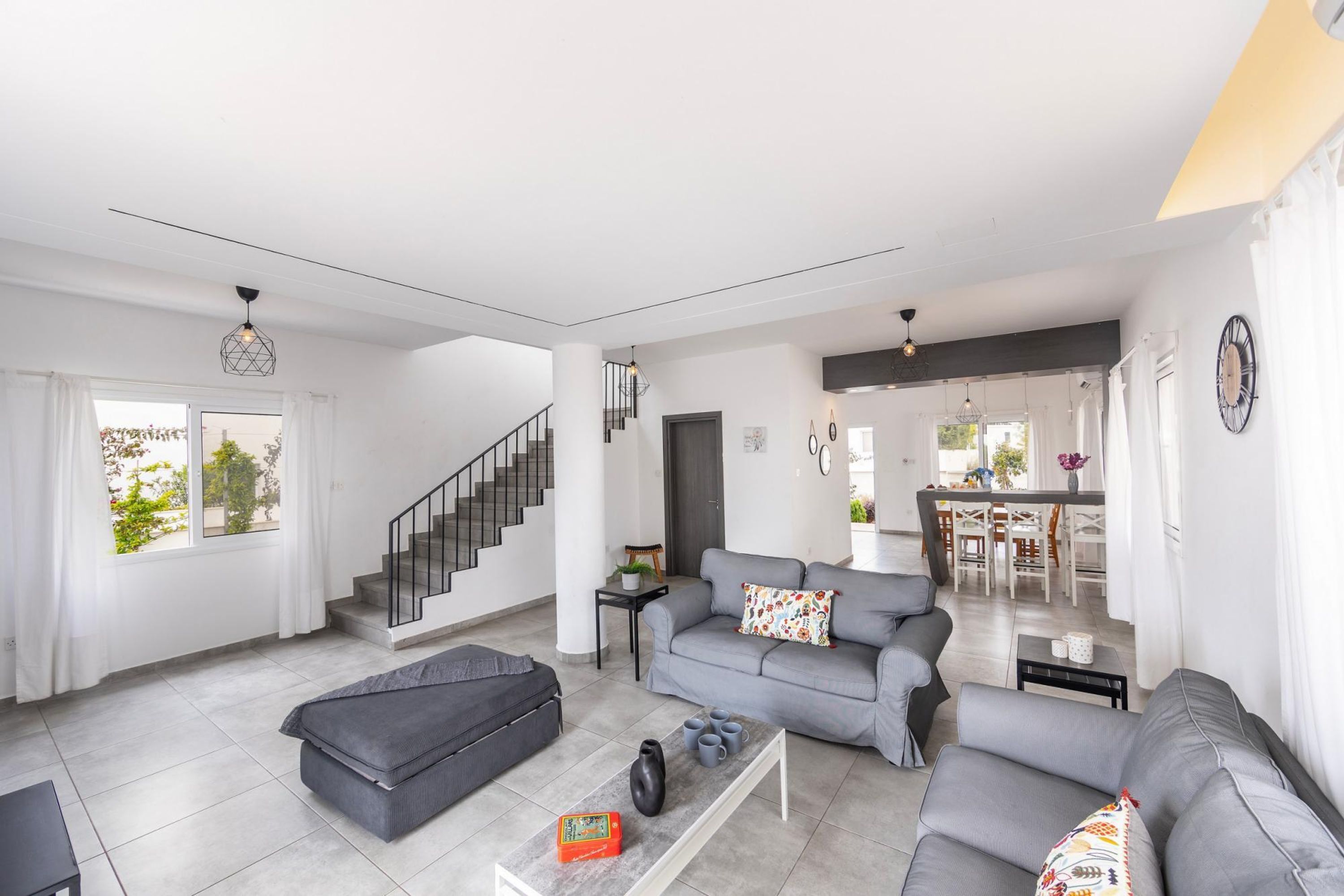 A modern living room with a grey sofa, a black ottoman, and a coffee table. There is a staircase leading to another level, and a dining area is visible in the background.