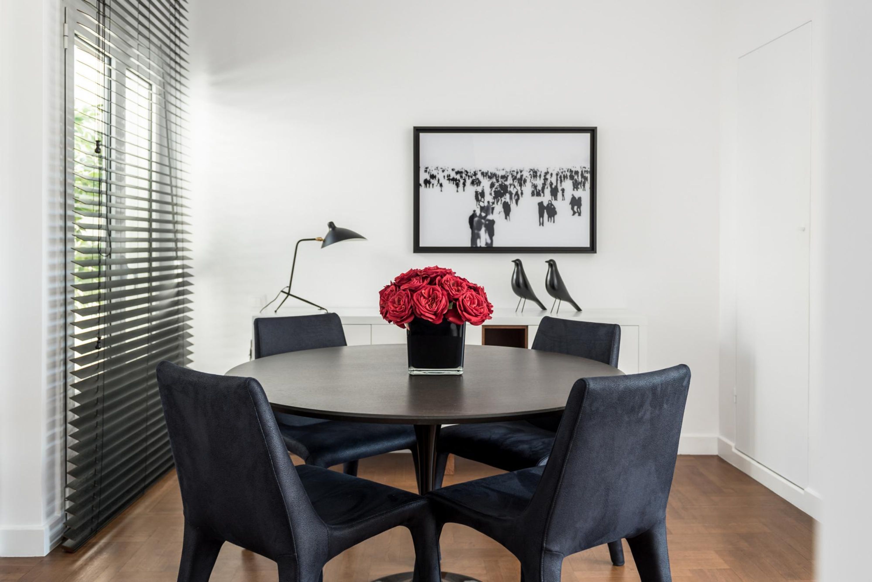A round dining table with six chairs, a vase of red flowers, a framed black and white photograph, and a black floor lamp.