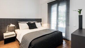 A modern bedroom with a black headboard, white bedding, a bedside table with a lamp, and a large window with blinds. - Executive 2 bdr Suite in Plaka - Acropolis View (Athens)