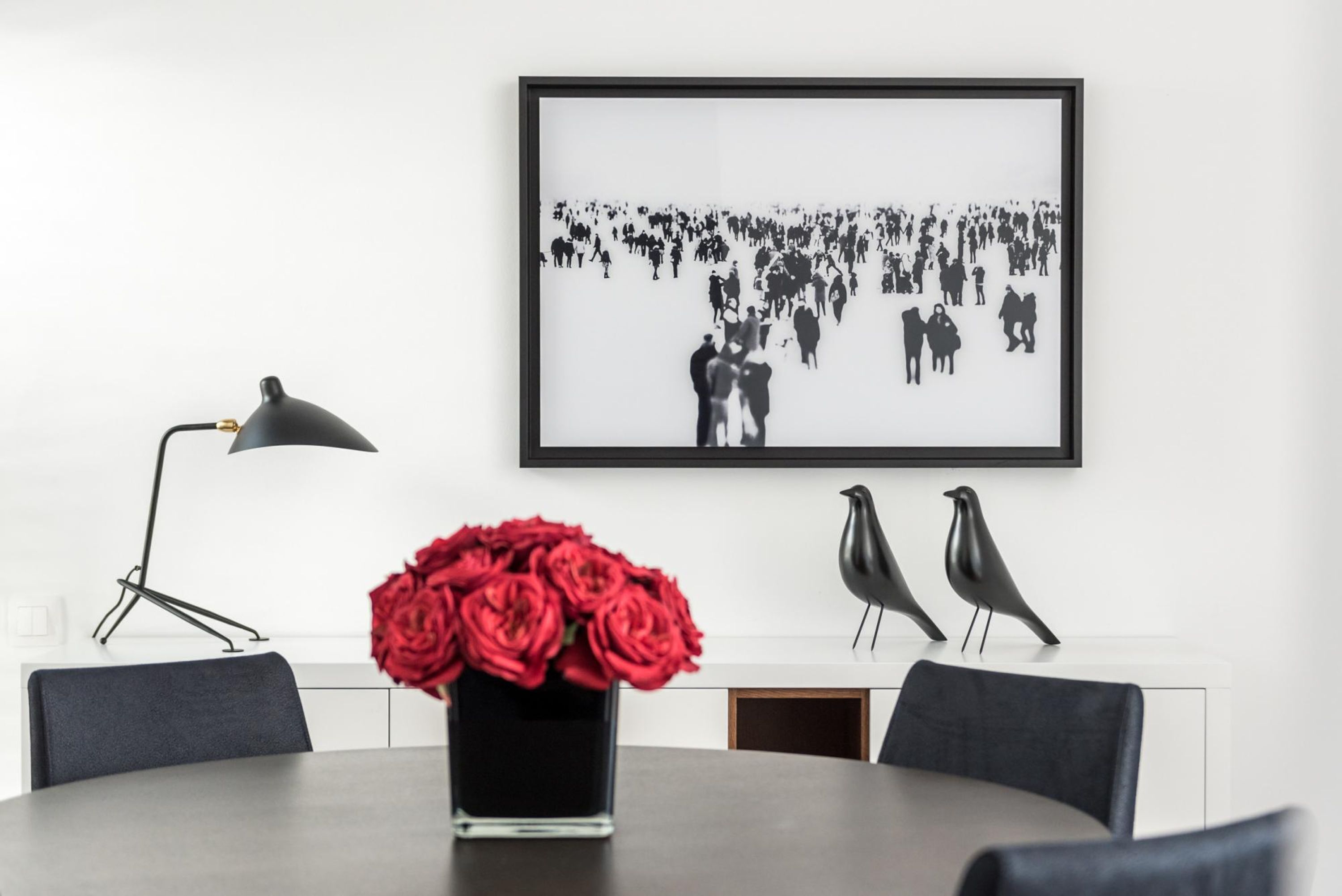 Een eetgedeelte met een ronde tafel, stoelen, een vaas rode rozen, een zwart-wit foto in een lijst, een vloerlamp en twee vogelbeeldjes.